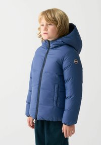 Giacca a vento blu con cappuccio, chiusura con zip e tasche laterali. Presenta un patch con il logo sulla manica. Texture liscia e design isolante.