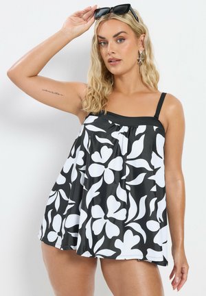 Mujer rubia con un traje de baño floral en blanco y negro y gafas de sol sobre la cabeza, levantando un brazo para ajustar las gafas de sol.