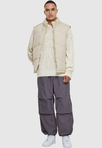 Beige pufferväst över en krämfärgad kabelstickad tröja, tillsammans med grå lösa byxor och vita sneakers, vilket ger en lager-på-lager, avslappnad look.