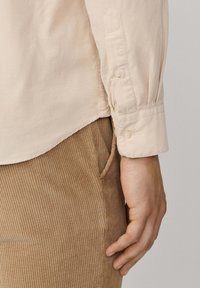 Chemise beige à manches longues avec des poignets à boutons, associée à un pantalon en velours côtelé marron texturé. La chemise a un ourlet incurvé et de légères plis.