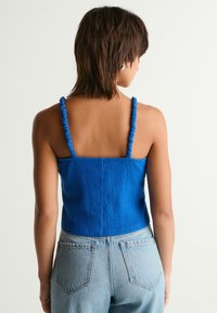 Top corto azul con tirantes fruncidos, presentado en una tela texturizada. Combinado con jeans de denim azul claro con bolsillos traseros.