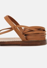 Sandale plate en cuir marron avec plusieurs fines lanières et semelle en bois sur fond blanc uni.