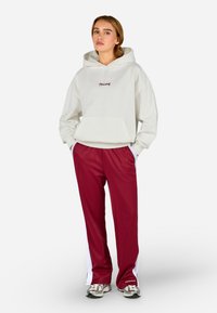 Kremowa bluza z kapturem z przednią kieszenią i logo 'TEGADOR'. Czerwone spodnie sportowe z białymi bocznymi paskami. Szare trampki z czarnymi akcentami.