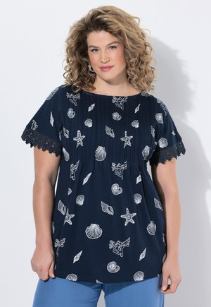 Frau mit lockigem Haar, die ein marineblaues Oberteil mit weißen Meereswesen- und Muschelmustern, spitzenbesetzte Ärmel und hellblaue Hosen trägt, vor weißem Hintergrund stehend.