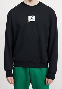 Sudadera negra con cuello redondo, mangas largas y un parche del logo de Jordan en blanco en el frente. Tela suave y texturizada; ajuste relajado.