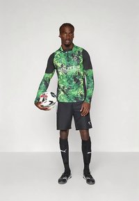 Grön och svart långärmad fotbolls-tröja med abstrakta mönster, matchad med svarta shorts och strumpor, håller en fotboll.