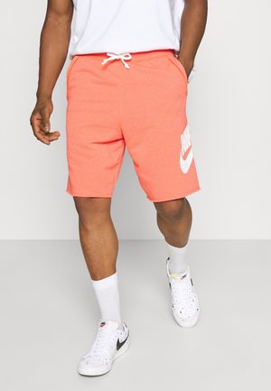 Homme portant un short Nike corail avec cordon blanc, des chaussettes Nike blanches et des baskets blanches, marchant devant un fond uni.