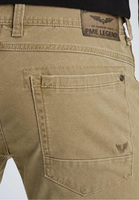 Beige cotton trousers with a textured finish, featuring a back pocket, stitched detailing, and a branded leather patch at the waistband.
