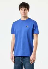 Blå bomulls t-skjorte med korte ermer, rund hals, og liten svart logo-patch på brystet, sammen med blå denim jeans.