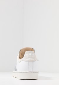 Zapatilla de cuero blanco con textura suave, interior de ante suave en tono marrón, rayas grises y suela texturizada blanca. Presenta un logo en relieve en el talón.