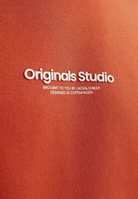 Oranje katoenstof met een gladde textuur; bevat witte reliëftekst: "Originals Studio," "Brought to you by JACK&JONES," en "Designed in Copenhagen."