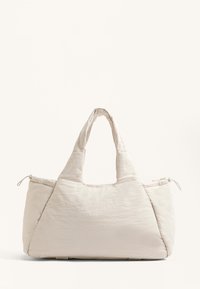 Beige stof tote bag med quiltet tekstur, brede håndtag og en afrundet form. Har en lynlåslukning på den ene side.