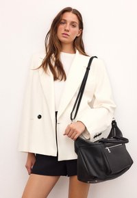 Fehér, oversized blézer fekete gombokkal, fekete táskával, amely zipzáras zsebbel és texturált bőrből készült. A fekete sortok teszik teljessé a megjelenést.