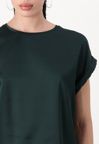 Mørkegrønn kortermet bluse med rund hals, laget av glatt stoff, med oppbrettede ermer og en avslappet passform.