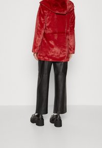 Rød falsk pelsjakke med hætte, der har en blød tekstur og en løs pasform. Båret over sorte læderbukser og chunky sorte sko.