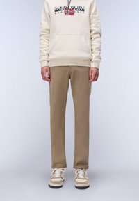 Beige hoodie med ficka fram och logotypdesign, ihop med ljusbruna slim-fit byxor och beige sneakers med vita snören och bruna accenter.