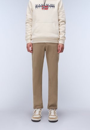 Beige Kapuzenpullover mit einer Vordertasche und Logo-Design, kombiniert mit hellbraunen Slim-Fit-Hosen und beigen Sneakers mit weißen Schnürsenkeln und braunen Akzenten.