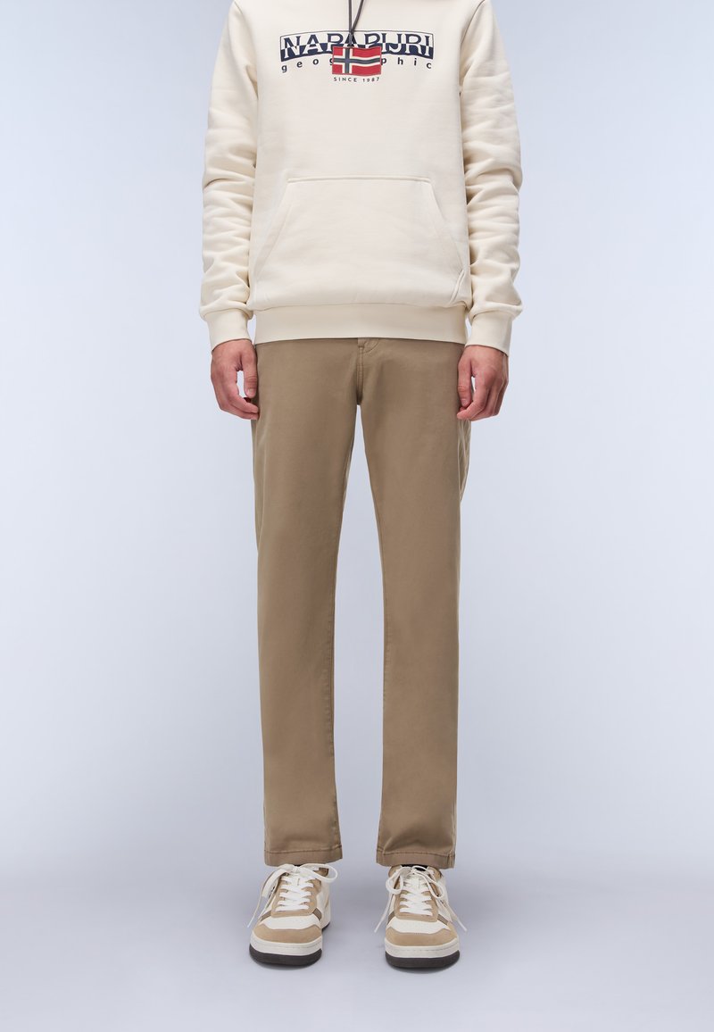 Beige hoodie med ficka fram och logotypdesign, ihop med ljusbruna slim-fit byxor och beige sneakers med vita snören och bruna accenter.