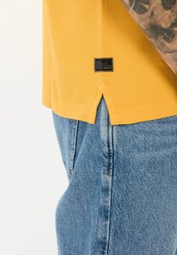 Geel T-shirt met korte mouwen van textiel met structuur en zijsplitten, gecombineerd met blauwe spijkerbroek. Voorzien van een zwart label aan de zoom.