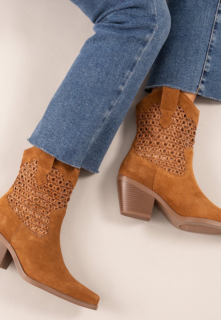 Botas de tobillo de ante marrón con un diseño de corte tejido en los costados, puntera puntiaguda y tacón apilado, combinadas con unos jeans de mezclilla azul.
