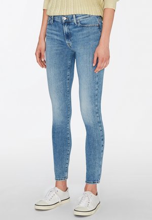 Persoon die lichtblauwe skinny jeans, een beige trui en witte sneakers draagt, staand tegen een effen witte achtergrond.