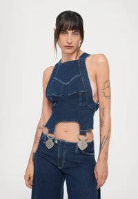 Tamni traper opušteni top s halter izrezom, isječenim stranama i srebrnim detaljima. Ima vidljive šavove i dizajn koji se tijesno prilagođava.