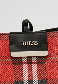 Étiquette en cuir noir avec le logo "GUESS" en argent sur un tissu à carreaux rouge et noir texturé, avec une petite boucle en cuir au-dessus de l'étiquette.