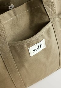 Plátěná taška v olivově zelené barvě s přední kapsou a etiketou s nápisem "self." Obsahuje detaily šití a pevné uši.