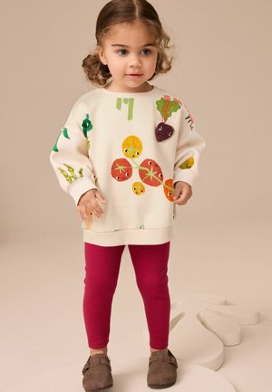 Cremefarbener Sweatshirt mit bunten Obst- und Gemüsemustern, kombiniert mit roten gerippten Leggings und braunen Schuhen.