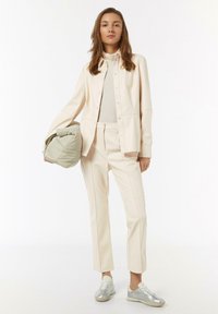 Cremfarbige Outfits mit einem Hemd mit Knöpfen und einer maßgeschneiderten Hose. Das Modell hält eine grüne Tasche und trägt silberne Sneakers mit einem eleganten Design.