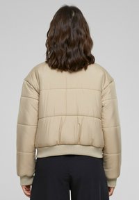 Beige vadderad jacka med ribbad nederkant, som har horisontella kviltade paneler och elastiska ärmslut. Slät yta, medellång silhuett.