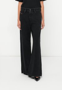 Μαύρα τζιν με φαρδύ πόδι και ψηλή μέση, με εμπρόσθιες τσέπες και διακριτική κόκκινη ραφή. Λείο, υφασμάτινο ύφασμα με υφή.