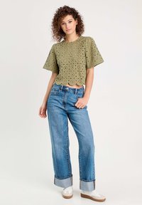 Crop top en dentelle olive avec des manches courtes et un ourlet en dentelle, associé à un jean bleu taille haute avec des revers exagérés et des baskets blanches.