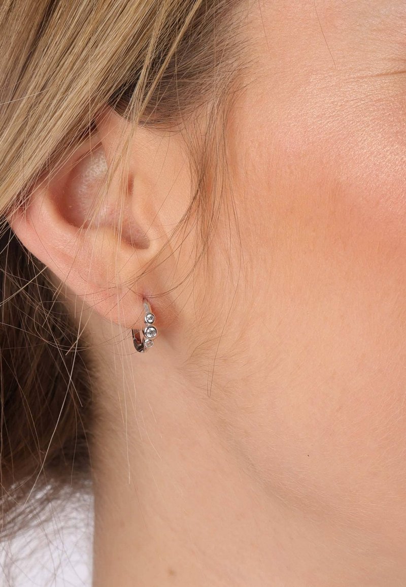 Boucles d'oreilles en argent en forme de cerceau avec des cristaux transparents, présentant un design épuré, une petite taille et une texture lisse, portées à l'oreille.