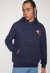 Sudadera azul marino con bolsillo tipo canguro y cordón. Presenta rosas bordadas en rojo y blanco en el pecho. Tejido suave y texturizado.