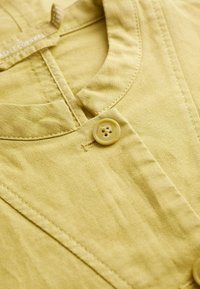 Tessuto di cotone giallo chiaro con colletto rotondo e chiusura con un solo bottone. Presenta cuciture visibili e una superficie texturizzata.