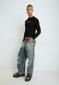 Schwarzes geripptes Langarmshirt mit silberner Stickerei; ausgebleichte hellblaue Schlagjeans mit Rissen; goldene und schwarze Plateau-Schuhe.