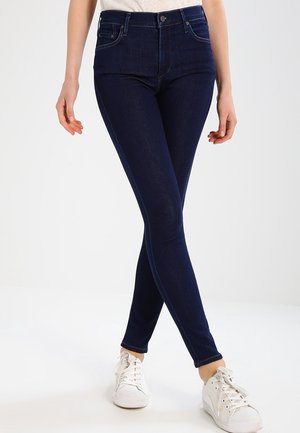 Femme portant un jean skinny bleu foncé et des baskets blanches, debout jambes croisées devant un fond blanc uni.