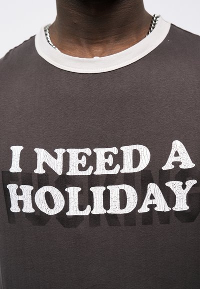 Άνδρας που φοράει σκούρο γκρι μπλουζάκι με λευκό γιακά και έντονο λευκό κείμενο μπροστά που γράφει "ΧΡΕΙΑΖΟΜΑΙ ΔΙΑΚΟΠΕΣ".