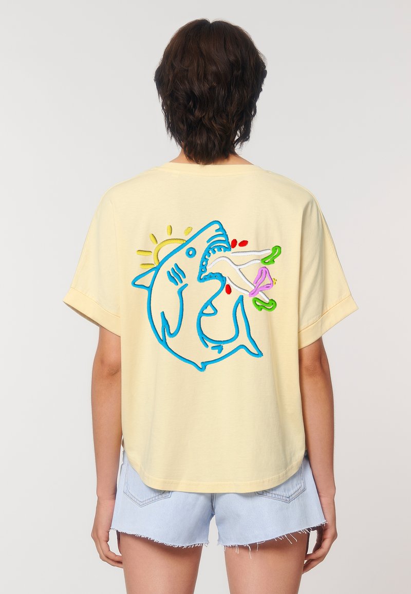 Geel katoenen t-shirt met een ontspannen pasvorm, voorzien van een blauwe geborduurde haai op de achterkant, die een gestileerde vis en zon vasthoudt.