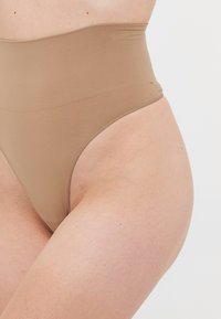 Intimo modellante beige a vita alta e senza cuciture indossato sui fianchi e nella parte alta delle cosce di una persona, su uno sfondo chiaro e neutro.