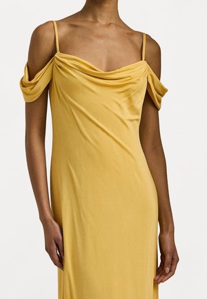 Femme portant une robe jaune en satin à épaules dénudées avec fines bretelles et tissu drapé sur les bras supérieurs sur un fond uni.