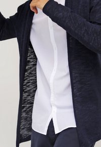 Námorně modrý cardigan na zapínání vyrobený z texturované pleteniny, vrstvený přes bílou halenu s knoflíky, který předvádí ležérní vzhled.