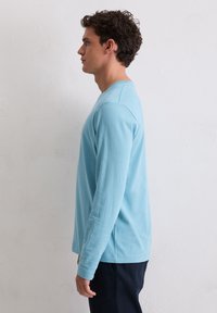 T-shirt met lange mouwen van lichtblauwe katoen, ronde hals, slim fit, met geribbelde manchetten en een gladde textuur, gedragen door een man vanaf de zijkant.