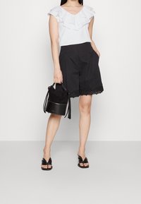 Blouse blanche à volants avec un col en V, short noir sur mesure avec bordure en dentelle, tenant un sac seau noir et portant des sandales noires.