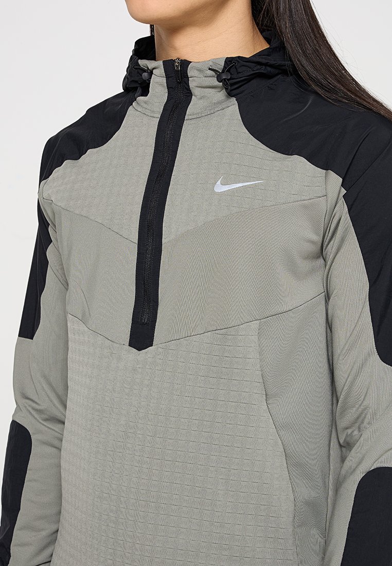 Veste à capuche Nike dans un design gris et noir, avec un zipper demi-long à l'avant, un tissu texturé et un cordon élastiqué au niveau de la capuche.