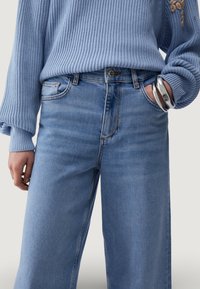 Hele sinine soonikkoes kampsun, millele on lisatud kõrge vöökohaga sinised teksad. Teksad on lõdva istuvusega ja neil on peened õmblused. Näha on hõbedased käevõrud.