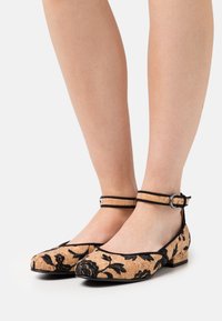 Dámské tan a černé květinové boty na nízkém podpatku s kotníkovými pásky, nošené na bosých nohou, zobrazené zepředu a z boku na bílém pozadí.