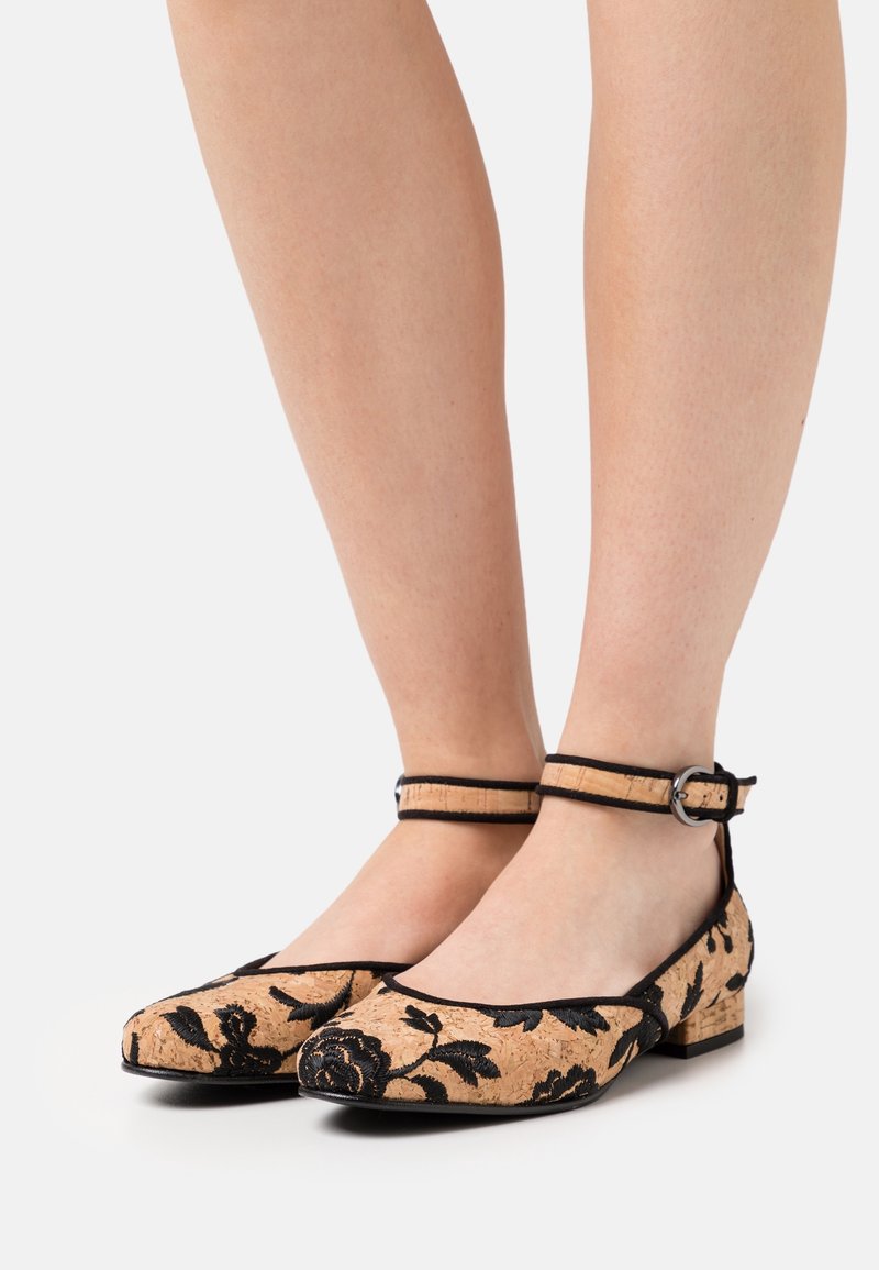 Dámské tan a černé květinové boty na nízkém podpatku s kotníkovými pásky, nošené na bosých nohou, zobrazené zepředu a z boku na bílém pozadí.