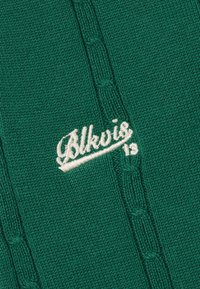 Tela de punto verde oscuro con texto "Blkvis 13" bordado en blanco y patrones verticales de punto trenzado.